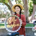 Woman holding a 'Hello Sunshine' sign at an outdoor market, wearing a straw hat and red sweater.