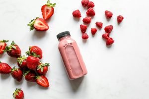 Top view of fresh strawberry and raspberry smoothie in a bottle with scattered fruits on white background.